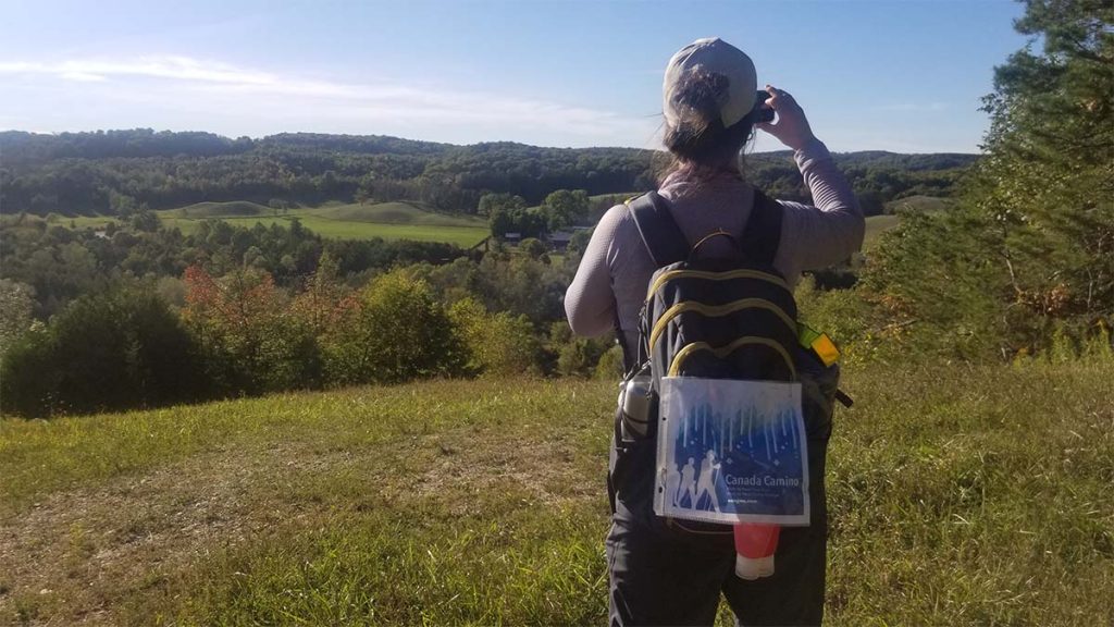Photo of a hiker wearing a backpack and a tag that reads "Canada Camino" They face away from the camera and are taking a photo of a landscape of trees and greenery.