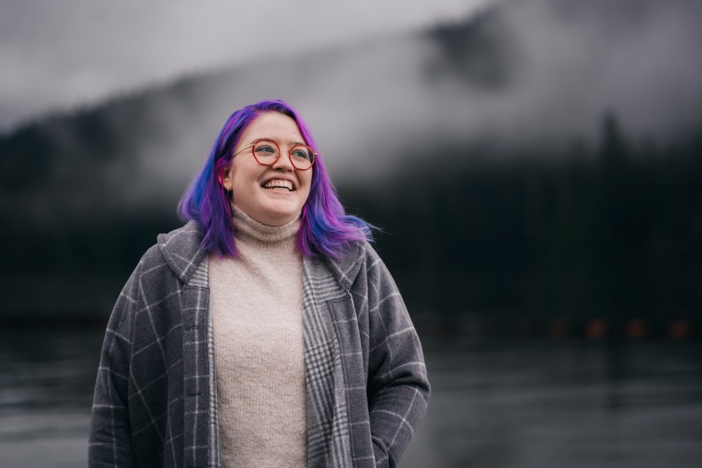 A photo of Caitlyn from the waist up in front of an out of focus background of water, forest and fog. She is laughing and looking off to the right.