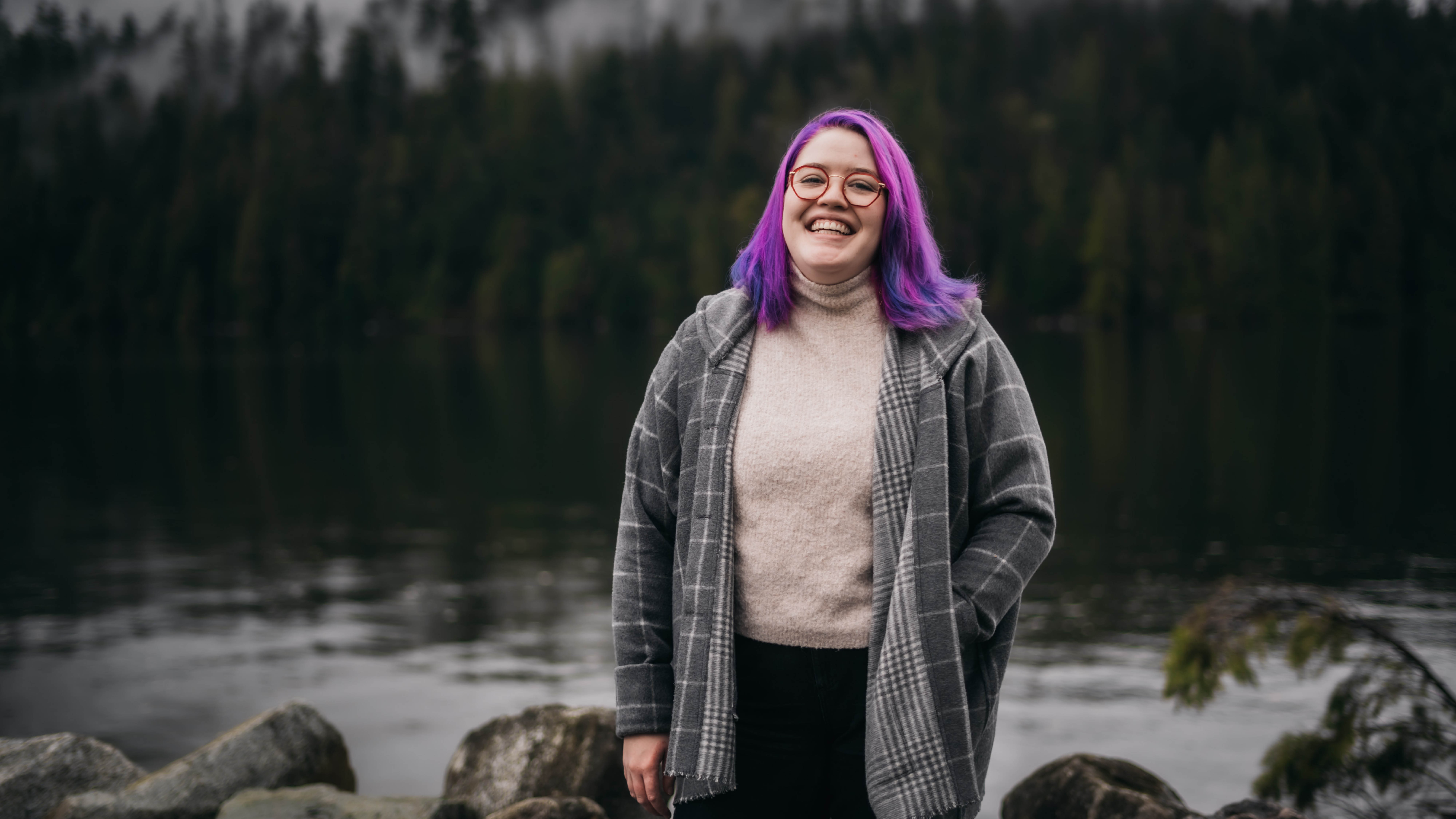 A photo of Caitlyn looking at the camera with a big smile. She is standing in front of large rocks and water. Across the water there is a green forest with clouds of fog dispersed throughout.