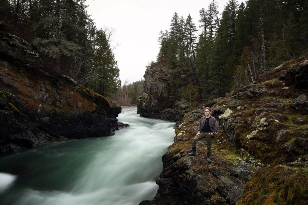 A photo of Levin standing next to a rushing river. He stands on a rock edge looking off into the distance.