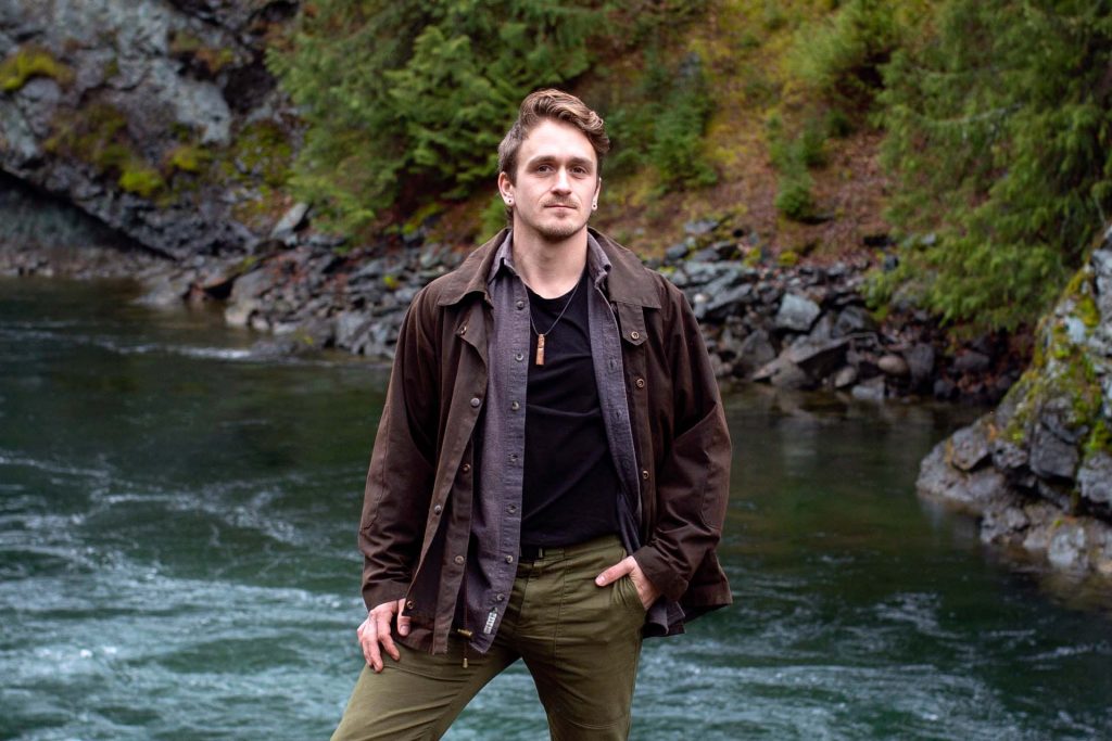 A photo of Levin standing in front of a river, looking at the camera. Moss covered rocks surround the river.