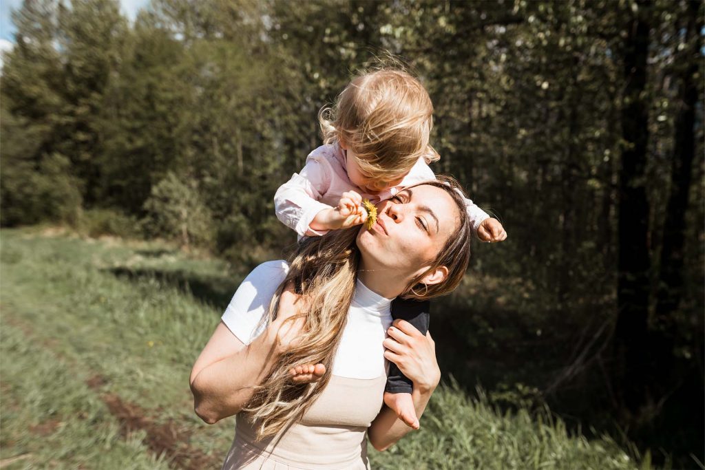 A photo of Jamie-Leigh and her kid, Rook. Rook is on her shoulders holding a dandelion up to her nose to smell. They are on a grassy path in front of a line of trees.