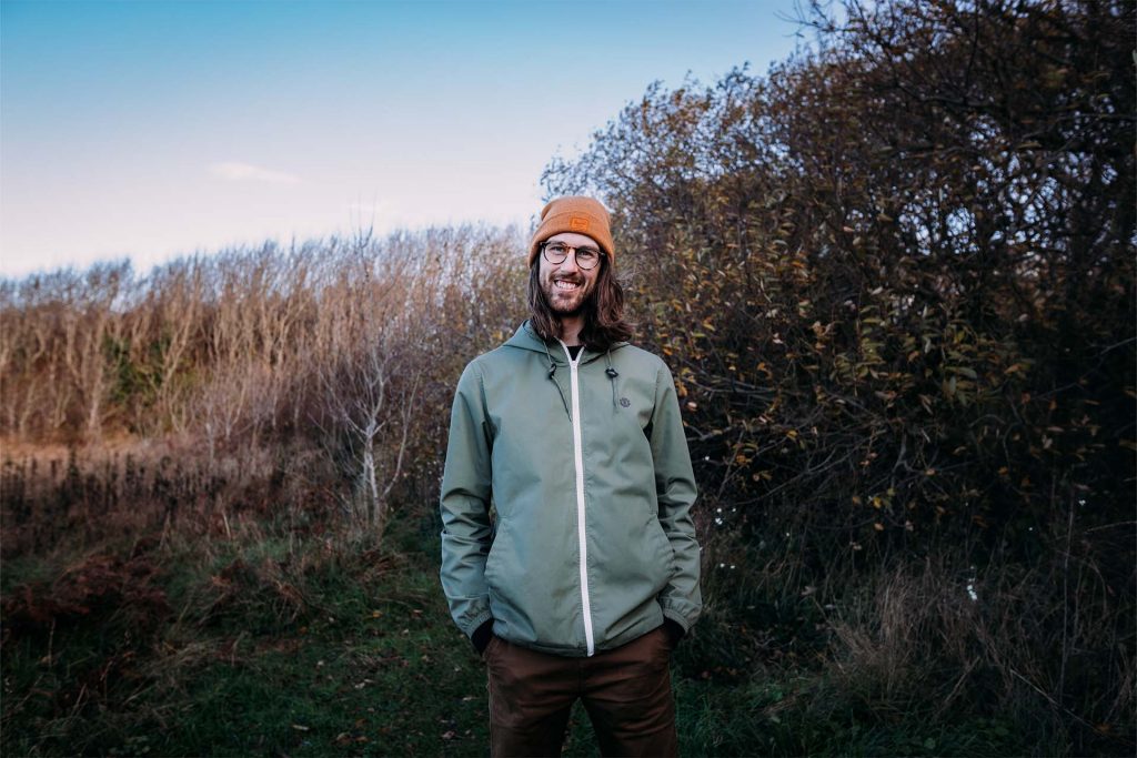 photo of russ standing with his hands in his pockets, smiling. He is surrounded by autumn trees that have shed their leaves.