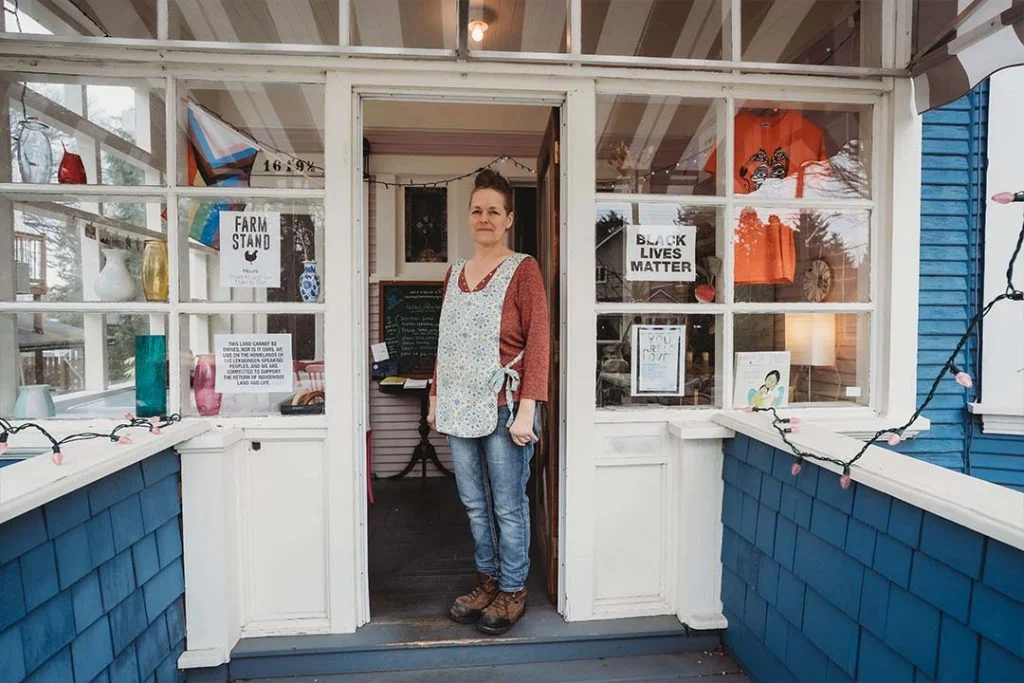 Marianne Unger stands in the doorway of her porch at the Farm Stand. The windows are filled with notes, and pictures and support for Black Lives Matter. Marianne is wearing a patterned apron over her clothes. 