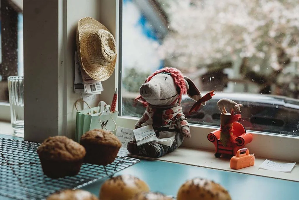 a small stuffed pig toy leans against a window with a hand written note resting in its lap. Muffins and other pastries are on cooling racks in the foreground.