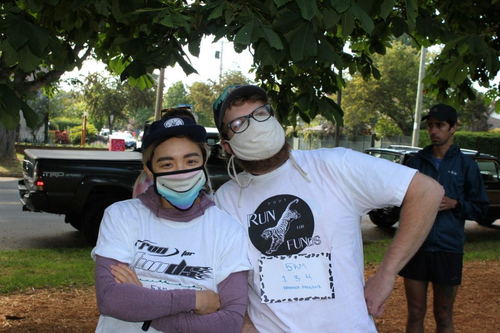 two people stand side by side wearing masks and seemingly smiling underneath them. They are outside with greenery and cars behind them. The person on the right is wearing a "Run for FUNds" shirt