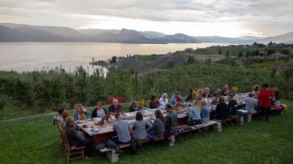 a wide shot of a long dinner table outside, filled with people. Mountains, hills and a lake are the backdrop.