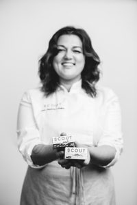Black and white photo of Chef Charlotte smiling and holding two containers of Scout’s canned seafood.