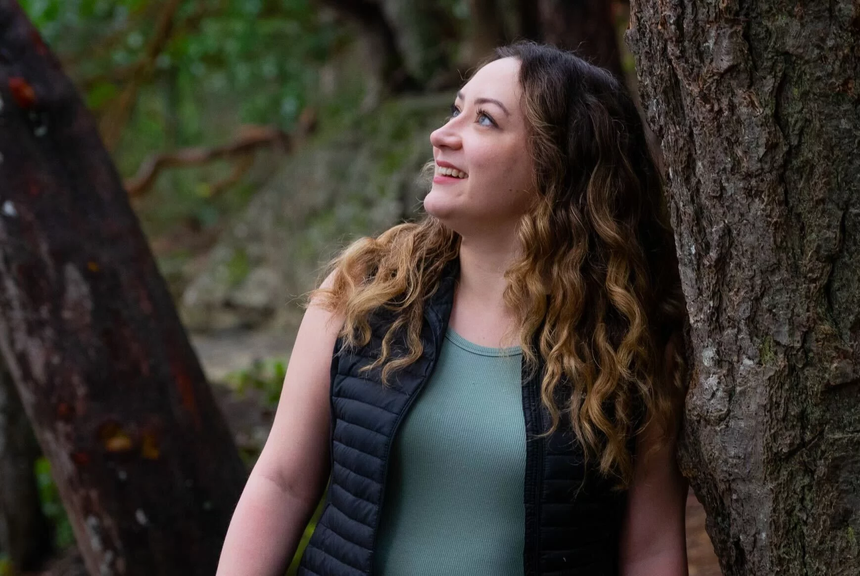 Mid shot of Carly looking up and to the side and leaning on a tree trunk.