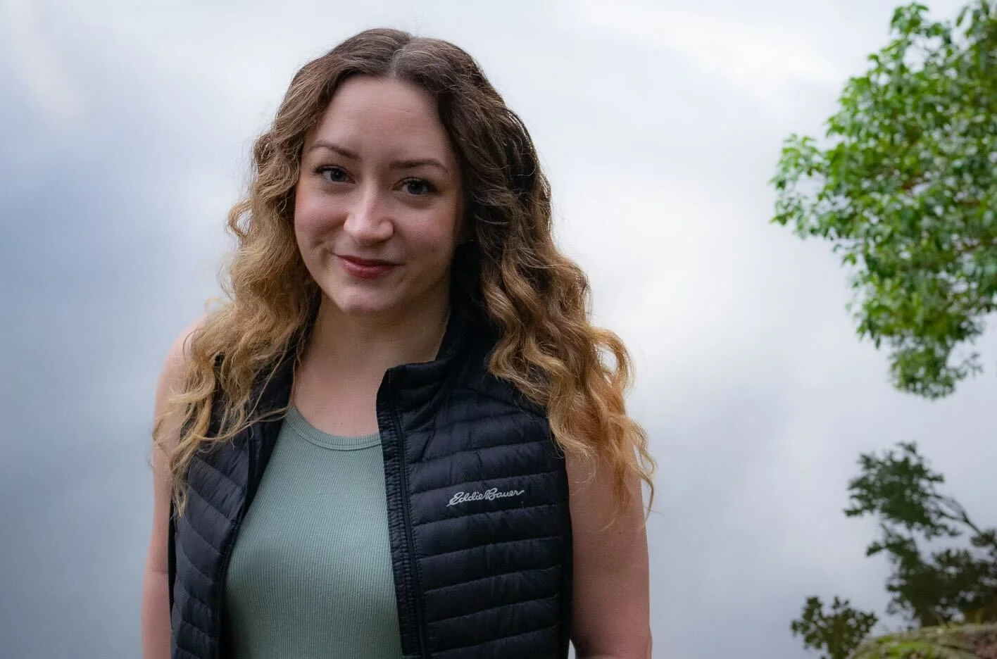 Photo of Carly from the bust up, looking at the camera with a smile. She stands in front of a cloudy/foggy background