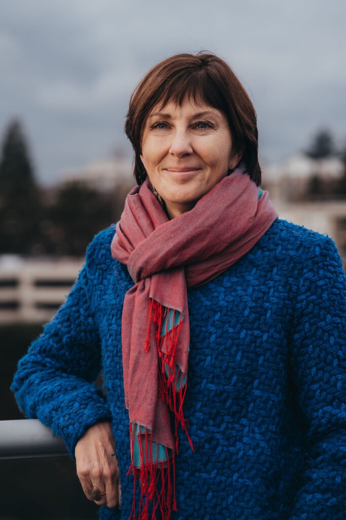 Photo of RAVEN's Executive Director, Susan Smitten, in a blue woolen coat and pink scarf, smiling at the camera knowingly.